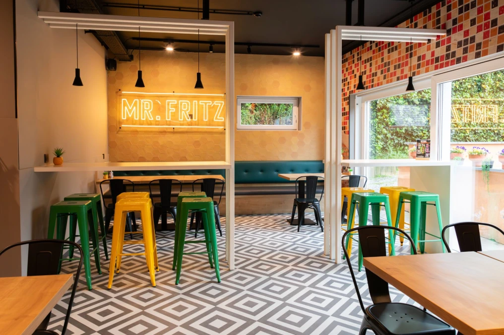 Entrée du snack : enseigne lumineuse, tabourets jaunes et verts, sol à losanges — Mr Fritz Fontaine