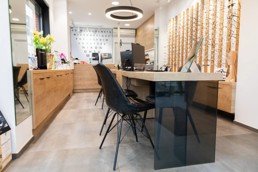 Espace conseil avec table sombre, chaises et présentoirs à lunettes en bois — Optique Wiard